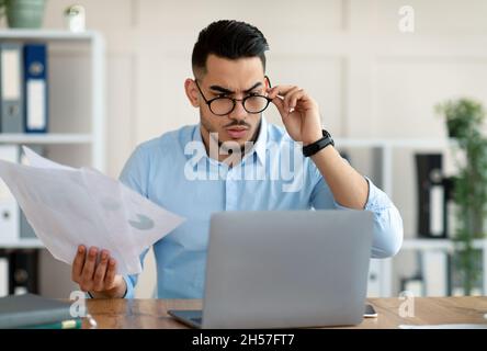 Stressed Arab employee with laptop and documents having problem with work, making mistake in business project at office Stock Photo