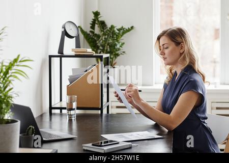Side view portrait of blonde businesswoman signing document while working at desk in home office, copy space Stock Photo