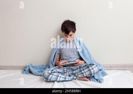 A brunette boy, a teenager in gray pajamas, on the floor, smiles, uses a digital tablet Stock Photo