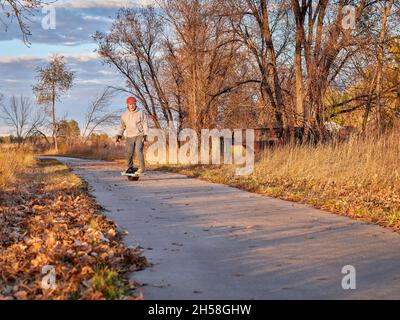 senior man is riding one-wheeled electric skateboard on a paved bike trail in fall scenery in Colorado Stock Photo