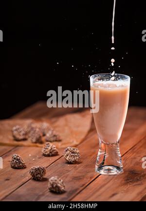 A stream of milk is poured into a glass with a milk drink with a splash of drops. Chocolate candies lie on a wooden table. Stock Photo