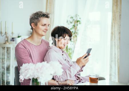 Portrait of happy couple man and woman using smartphone while sitting at the table together during breakfast in the kitchen at home. Scrolling social Stock Photo