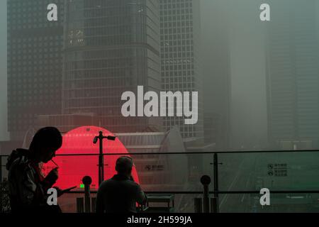 A young man lights a cigarette while another looks at the skyscrapers shrouded in severe smog in Beijing CBD, China. 06-Nov-2021 Stock Photo