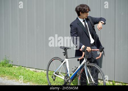 Asian young businessman walking outdoors and pushing a bicycle while looking at his wristwatch. Stock Photo