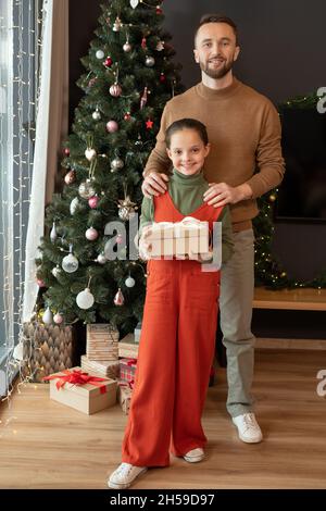 Portrait of happy middle-aged father in sweater holding hands on daughters shoulder against Christmas tree at home Stock Photo