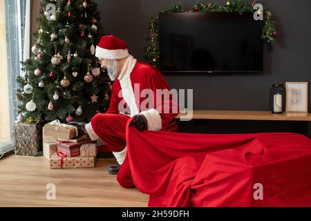 Gray-bearded Santa Claus in cap crouching at Christmas tree and putting gifts under it in living room Stock Photo