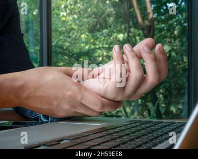 Hand pain from using a computer. Close-up man holding his wrist near laptop on green nature background, working at home. Office syndrome hand pain by Stock Photo