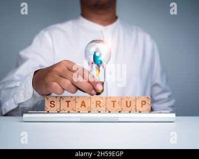 Business starts up with creative thinking for leadership concept. The rocket in idea light bulb holding by businessman and words 'STARTUP' on wood cub Stock Photo