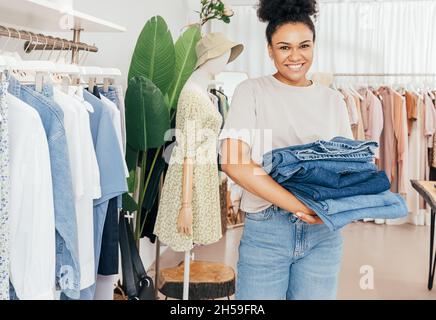 Smiling female fashion store worker holding a pile of jeans and looking at camera Stock Photo