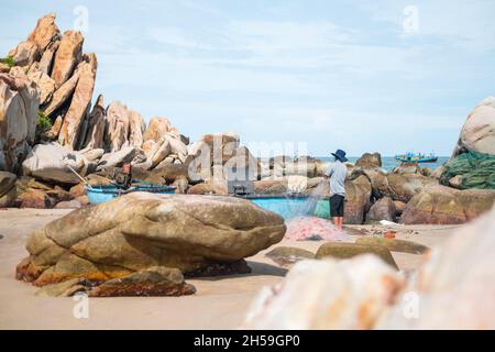 Local fisherman in the hat rolls up fish nets on the seashore. Picturesque beach with large rocks. Amazing places in the world. Real people life.  Stock Photo