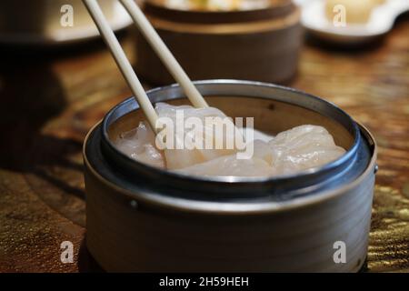 A close-up picture of bamboo chopsticks picking up white white Chinese stuffed steamed dumpling, also known as dim sum from a round bamboo basket on a Stock Photo