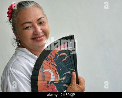 Smiling elegant mature Mexican Latina woman with red flower in grey hair holds handheld Asian fan and looks at viewer in front of grey wall. Stock Photo