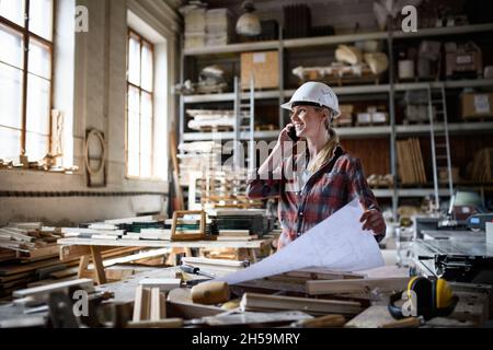 Female engineer holding blueprints and making phone call indoors in carpentry workshop. Stock Photo
