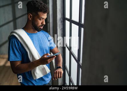 Young African American sportsman standing indoors at gym, using smartphone and smartwatch. Stock Photo