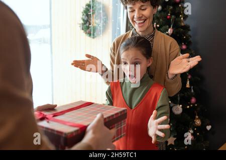 Amazed girl throwing hands up and keeping mouth open while getting Christmas present from parents Stock Photo