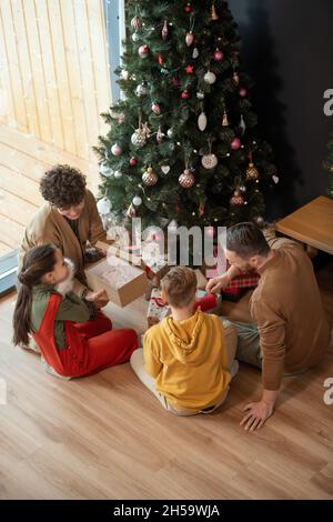 Parents sitting at decorated tree and gift boxes under it and giving Christmas Presents to children Stock Photo