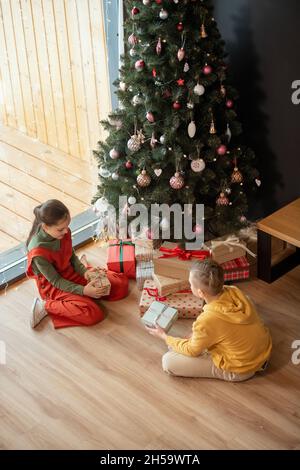 Siblings in bright outfits sitting with gift boxes on floor at Christmas tree and guessing what is inside of boxes Stock Photo