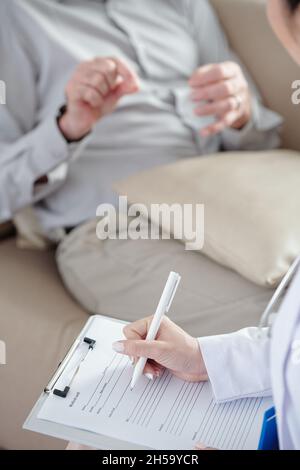 Hands of doctor obtaining and writing down anamnesis of senior patient when talking to him Stock Photo