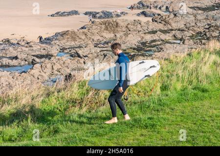 A surfer carrying his surfboard walking down a footpath to Fistral Beach at low tide in Newquay in Cornwall. Stock Photo
