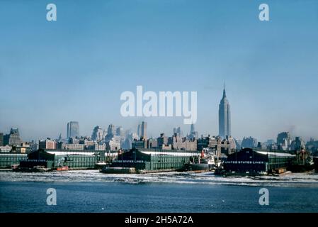 New York City, USA – an atmospheric view in the winter sunshine looking over the city from the Hudson River and the city’s docks in the mid-1950s. Ships in port are moored by the piers/jetties. The warehouses are those of the United States Lines. Ice is evident in the water. In the background a relatively low-rise skyline is dominated by the Empire State Building. This image is from an old Kodak colour transparency taken by an amateur photographer – a vintage 1950s photograph. Stock Photo