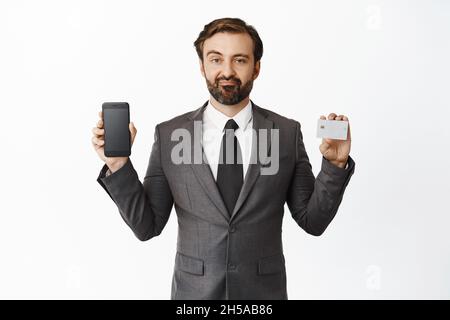 Skeptical man in suit shows his credit card and smartphone screen, smirk and look disappointed, negative opinion about phone app, white background Stock Photo