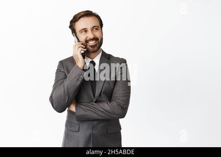 Handsome realtor smiling, talking on mobile phone and looking at upper right corner empty space, banner, standing over white background Stock Photo