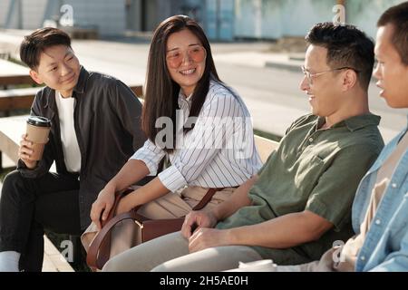 Group of young Chinese employees in casualwear sitting on bench and interacting in urban environment Stock Photo