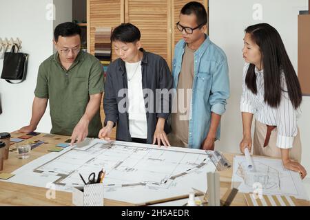 Group of young architects gathered by table with large sketch on blueprint discussing working points at meeting Stock Photo