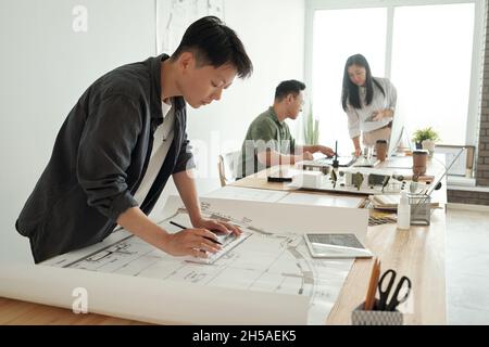 Young female architect or engineer drawing sketch while bending over table against her two co-workers in large openspace office Stock Photo