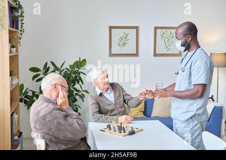 Male doctor giving the glass of water and pills to senior man while he sitting at the table and playing chess with his friend Stock Photo