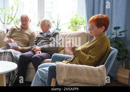 Senior woman sitting in armchair and knitting with two senior men discussing photos together on sofa in the background Stock Photo