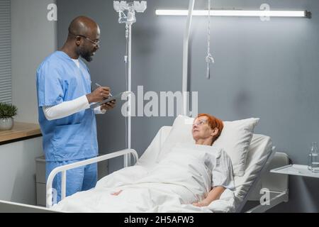 African male doctor writing prescription for his senior patient while she lying on the bed in the hospital ward Stock Photo