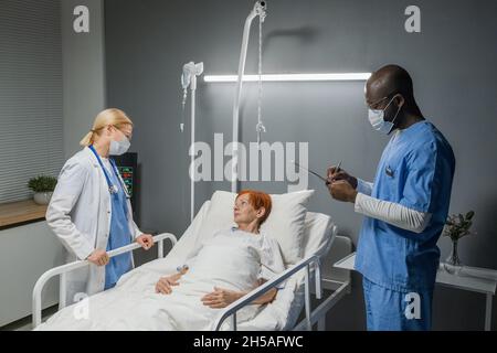 Two doctors standing near the bed with senior woman and learning about her health in the hospital ward Stock Photo