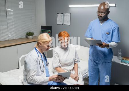 Female doctor showing something on tablet pc to senior woman while they sitting on the bed in ward with nurse making notes in medical card Stock Photo