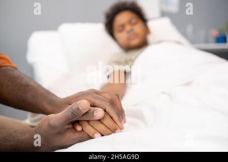 Close-up of father holding hand of his sick little son while he lying in bed at hospital Stock Photo