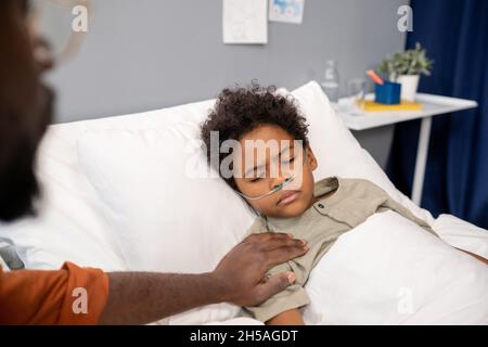Sick little boy lying in bed with his eyes closed while father sitting near the bed and supporting him Stock Photo