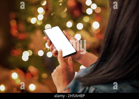 Christmas phone mockup in woman hand. Sending Christmas greetings by phone concept. Bokeh lights of decorated Christmas tree in background Stock Photo
