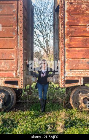 Adult blonde woman with sunglasses standing between two abandoned train cars Stock Photo