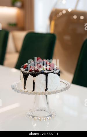 Gourmet cake with chocolate topping and berries on a glass stand on a white table in a modern interior. Soft selective focus. Stock Photo