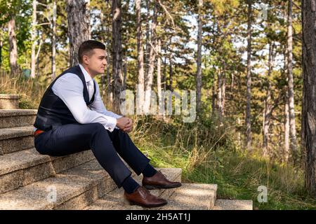 Handsome young groom wearing elegant and stylish dark blue suit outdoors portrait. Stylish groom sitting on stairs side view. Stock Photo