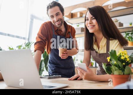Positive sellers in aprons holding smartphone and paper cup in flower shop Stock Photo