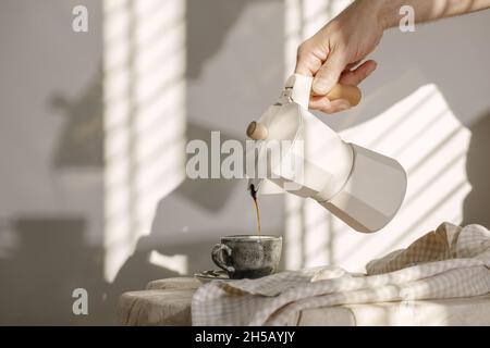 Pouring coffee from Italian coffee maker with wooden handle into grey espresso craft cup on solid wood stump decorated with linen tea towel in creamy