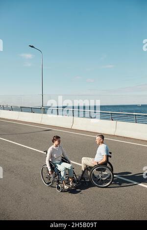Vertical portrait of adult couple in wheelchairs chatting outdoors and enjoying city life, copy space Stock Photo