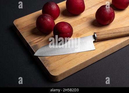 Red plums on bread board with knife and black background Stock Photo