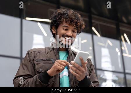 Happy young Indian man smiling holding mobile phone text social media outdoors Stock Photo