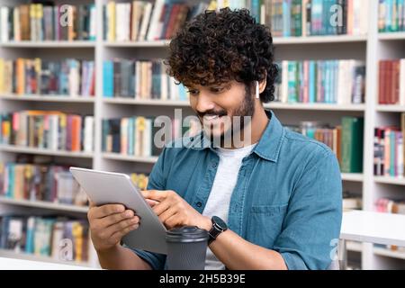 Young indian male student wearing earphones use mobile tablet sitting in library Stock Photo