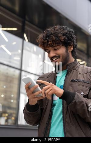 Happy smiling young Indian man walking city street using mobile phone  Stock Photo