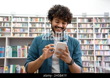 Overjoyed young Indian student chatting social media on smartphone in library Stock Photo