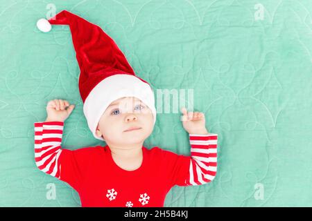 Smiling toddler boy lying on green coverlet. Christmas baby wearing big red Santa hat looking at camera. New Year concept. Top view. Copy space Stock Photo