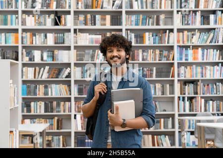Young smiling Indian male student standing in university library with backpack Stock Photo